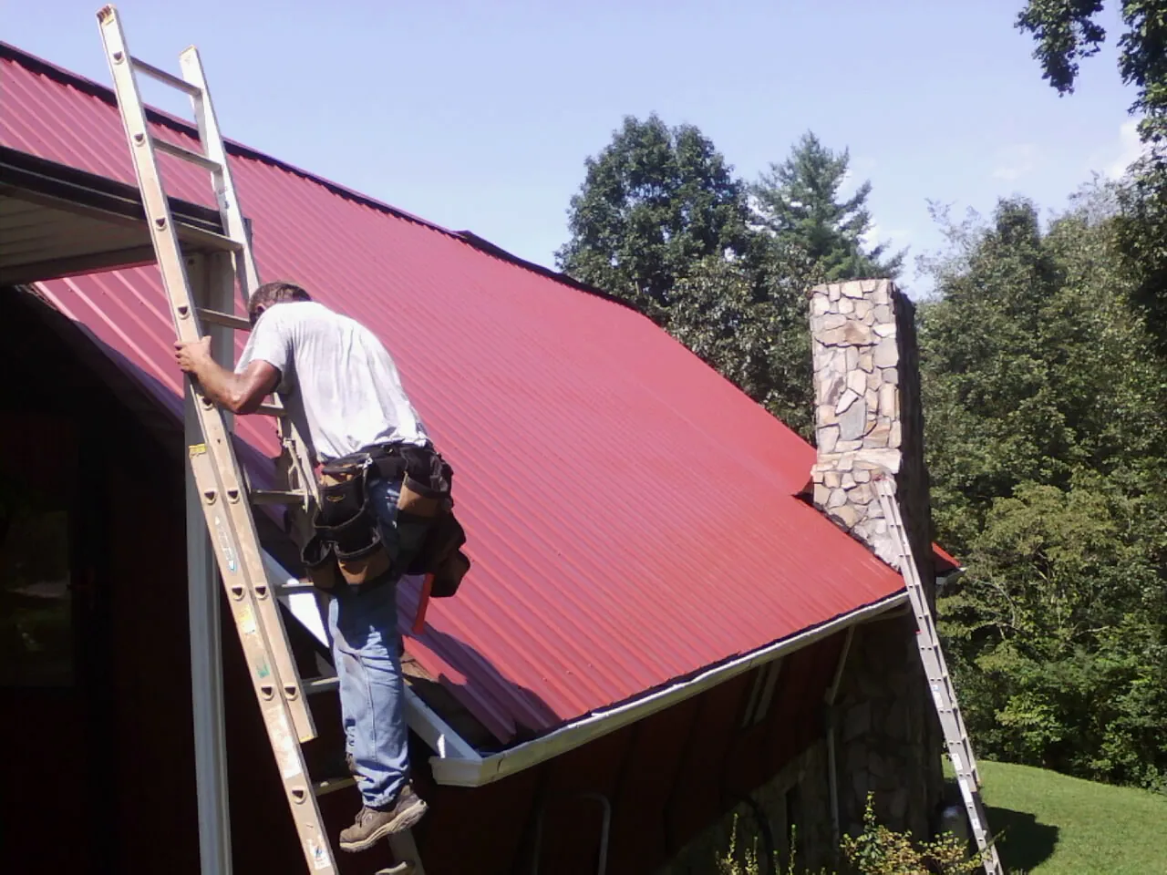 Roofer on a ladder beside a red metal roof and stone chimney — Soffit & Fascia Repair by Roofing Brothers in Withams